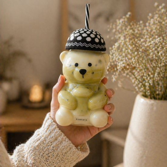 Person holding a bear-shaped green drink container with a straw, set against a blurred indoor background.
