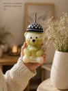 Person holding a bear-shaped green drink container with a straw, set against a blurred indoor background.